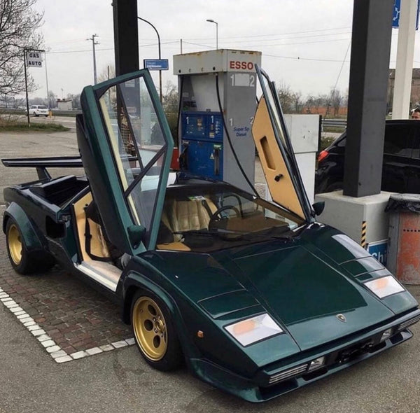 Lamborghini Countach in green with gold wheels and scissor doors open at gas station.